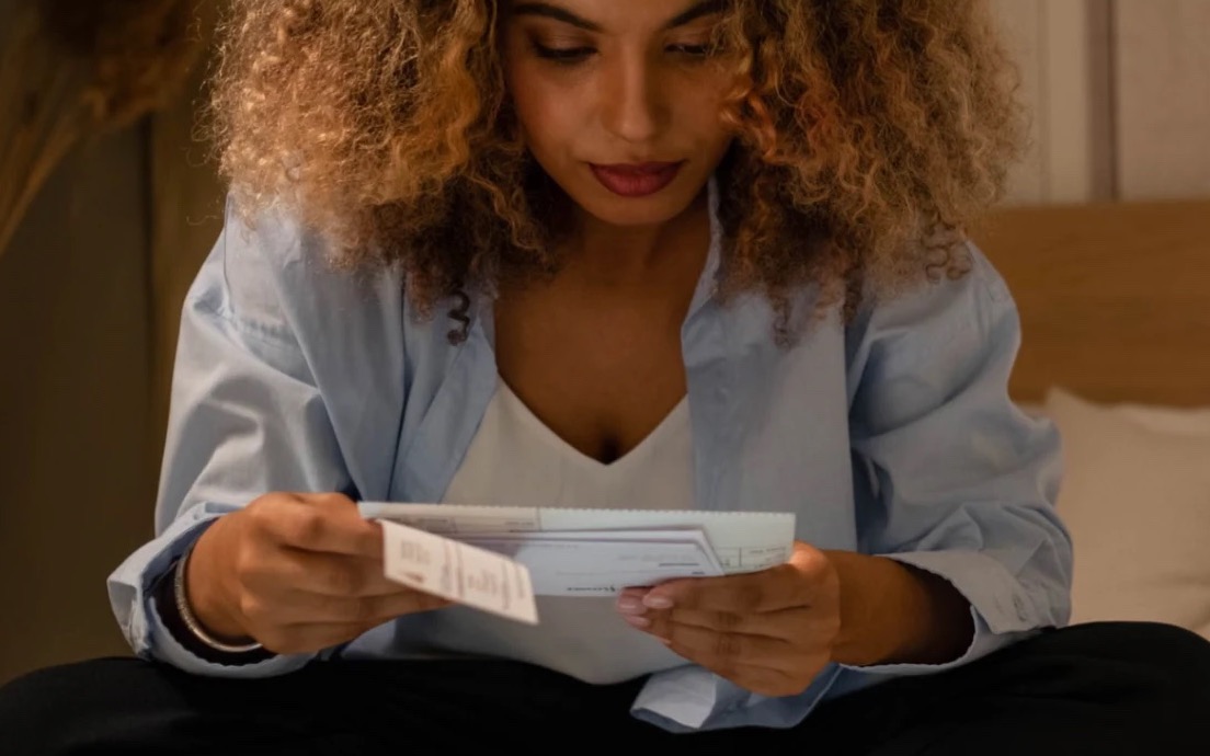 A woman sitting on her bed at night, reading through a bill